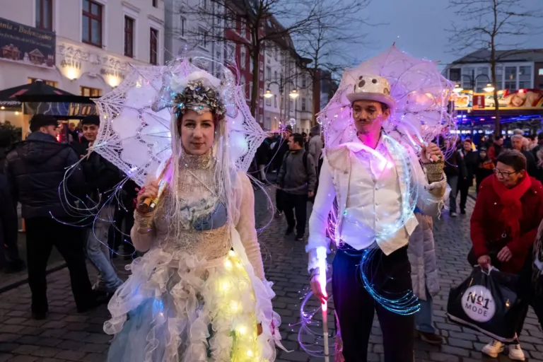 Zwei Kostümierte in weißen, beleuchteten Fantasiegewändern mit Spitzenschirmen in der Innenstadt